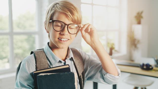 Boy in glasses smiles and holds books in classroom.
