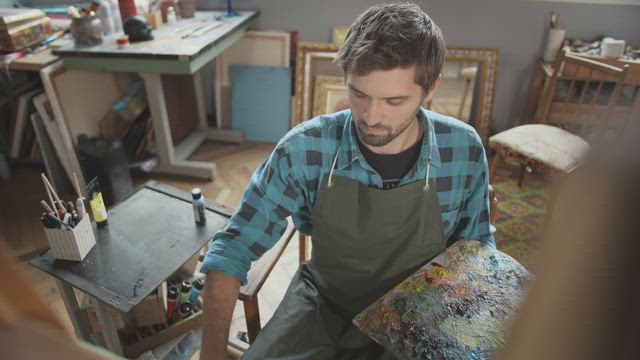 Artist with palette thinks about ideas while looking at canvas.